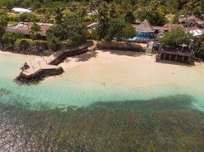 Vista aérea de un complejo turístico en la playa con aguas turquesas cristalinas, una playa de arena y una zona de piscina con sombrillas.