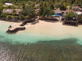 An aerial view of a beach resort with clear turquoise waters, a sandy beach, and a pool area with umbrellas.