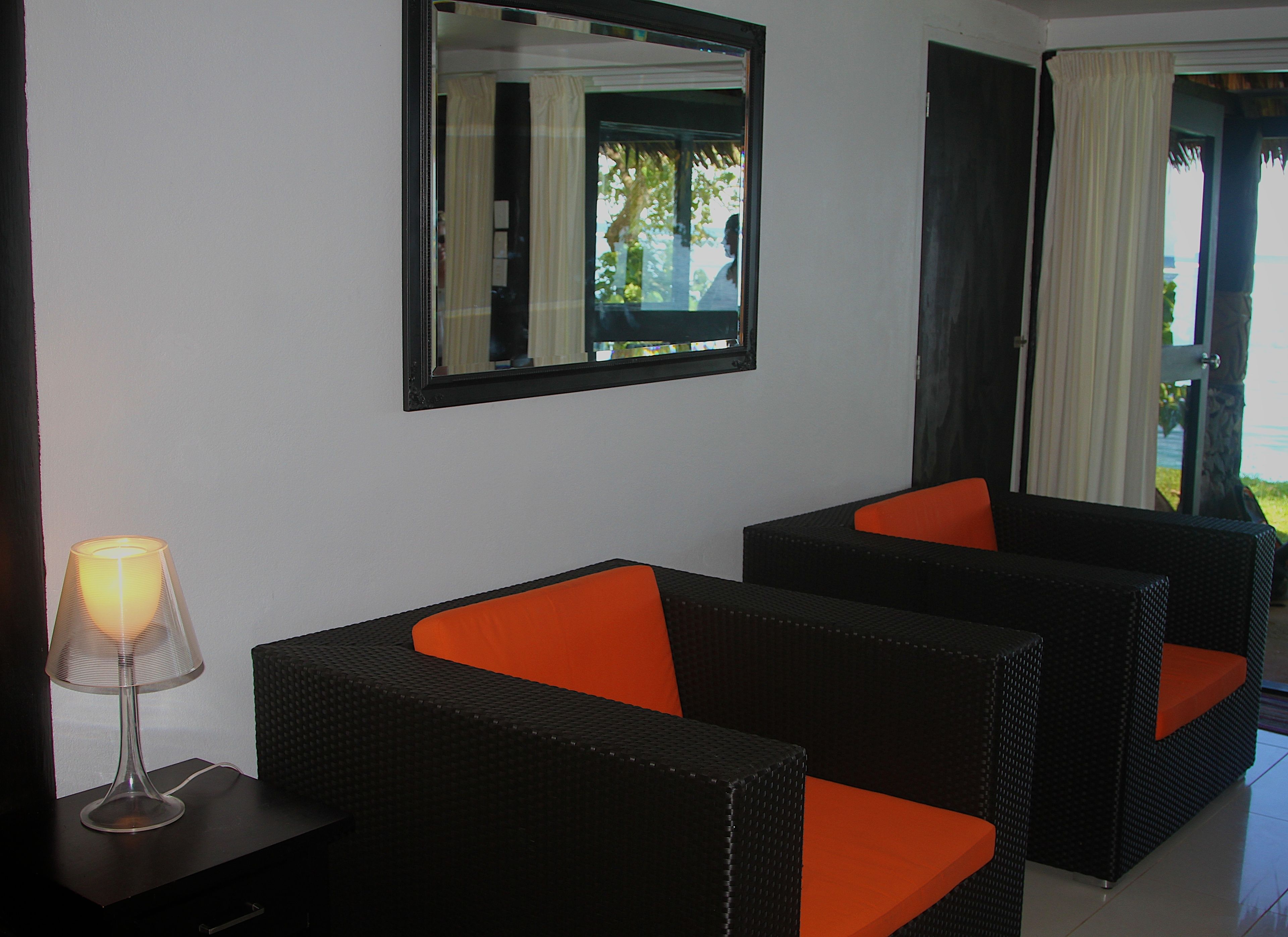 A room with two black wicker sofas with orange cushions, a small table with a lamp, a mirror on the wall, and a door leading to an outdoor area.