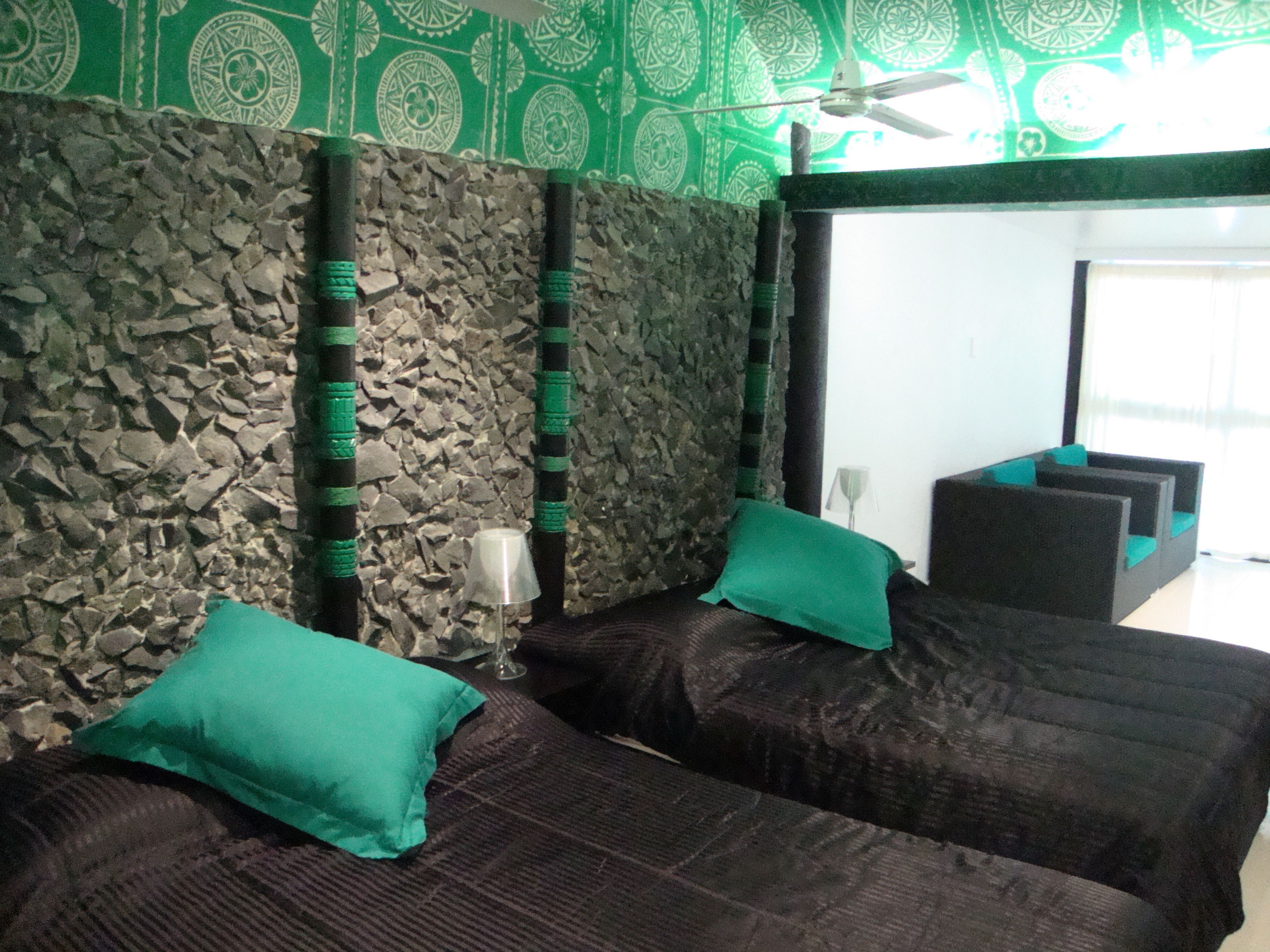 A room with a stone wall, two dark beds with green pillows, a ceiling fan, and a large mirror reflecting a seating area.
