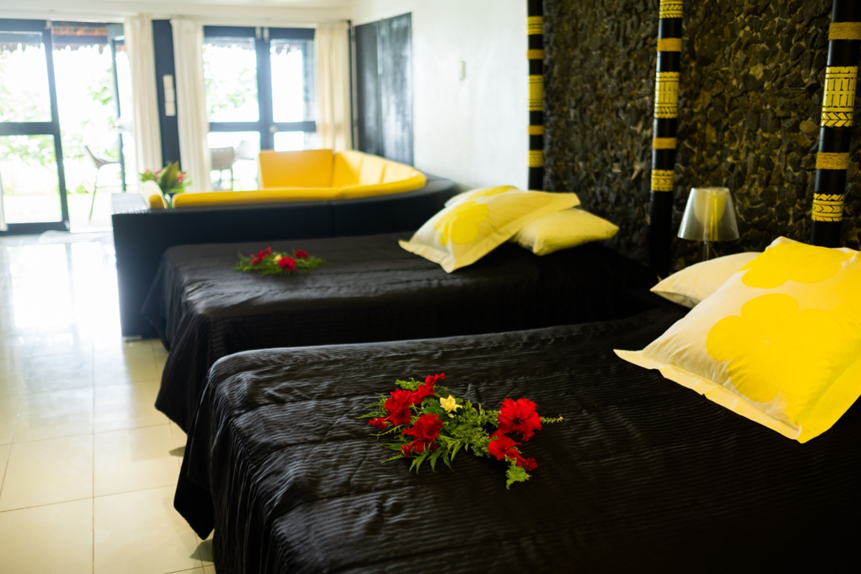 A hotel room with two beds, a yellow chair, a small table, and a window with curtains.