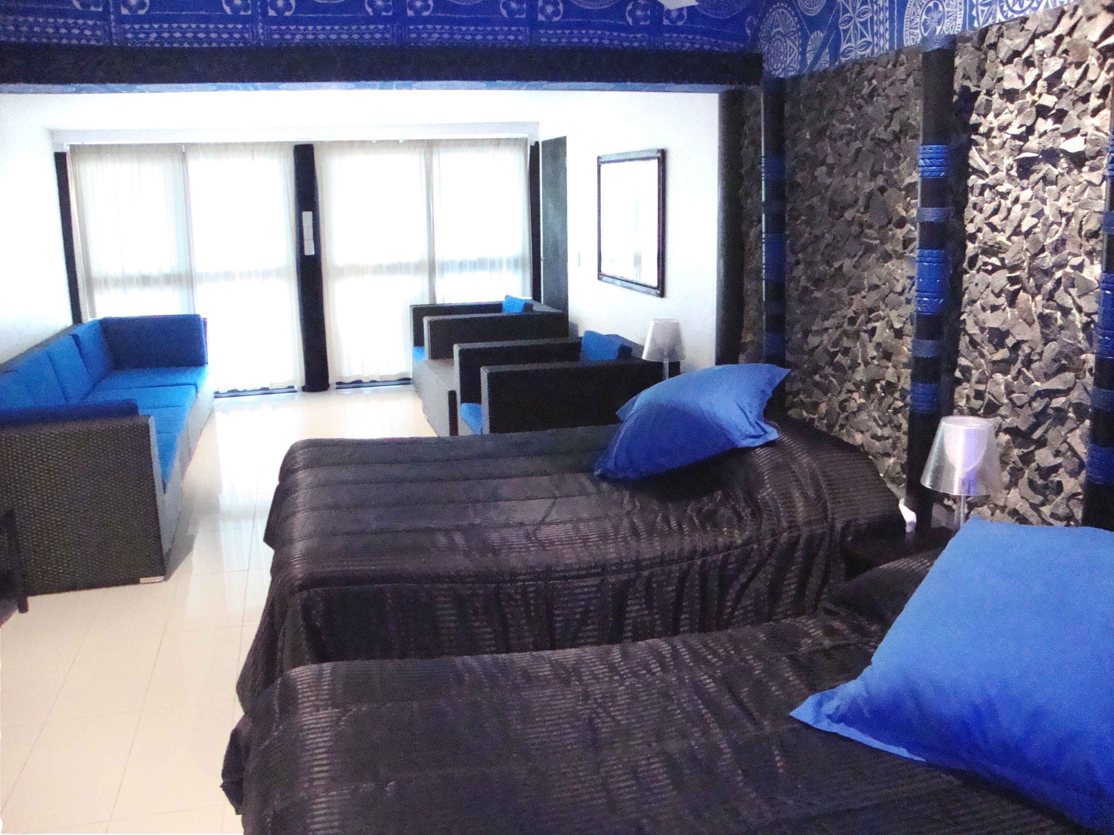 A hotel room with a bed, a sofa, and a chair. There is a window with curtains, a wall with a patterned design, and a door.
