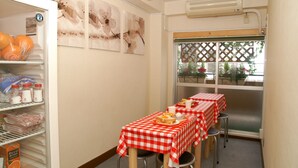 A compact room with a dining area featuring two tables with red and white checkered tablecloths, a refrigerator stocked with food items, and a window with a view of greenery outside. - Hostel Zen (Yokohama)