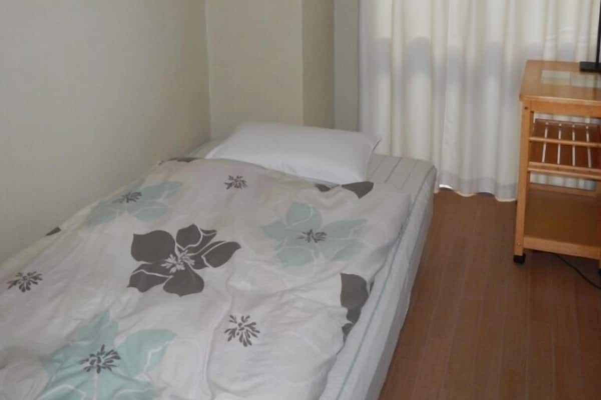 A small, single-bed room with a wooden nightstand, white curtains, and a floral bedspread.