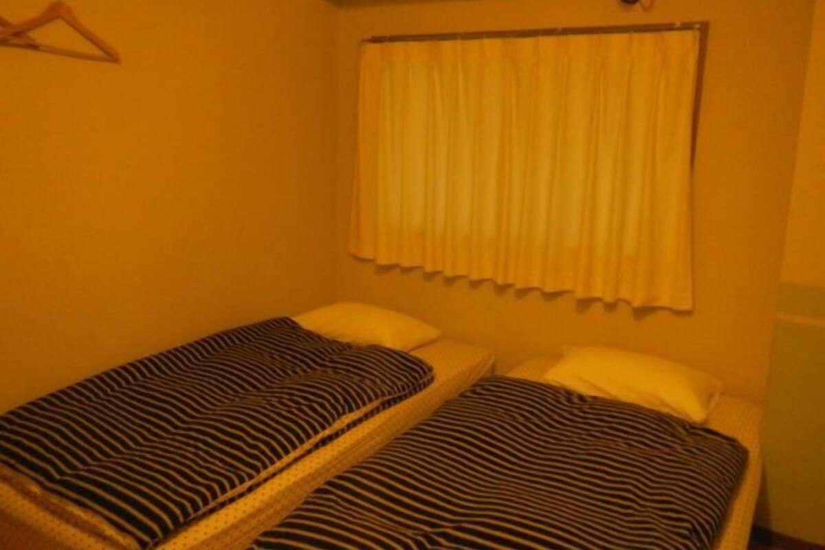 A small room with two single beds, each with striped bedding. There is a window with a curtain, and a wall-mounted air conditioning unit above the beds.