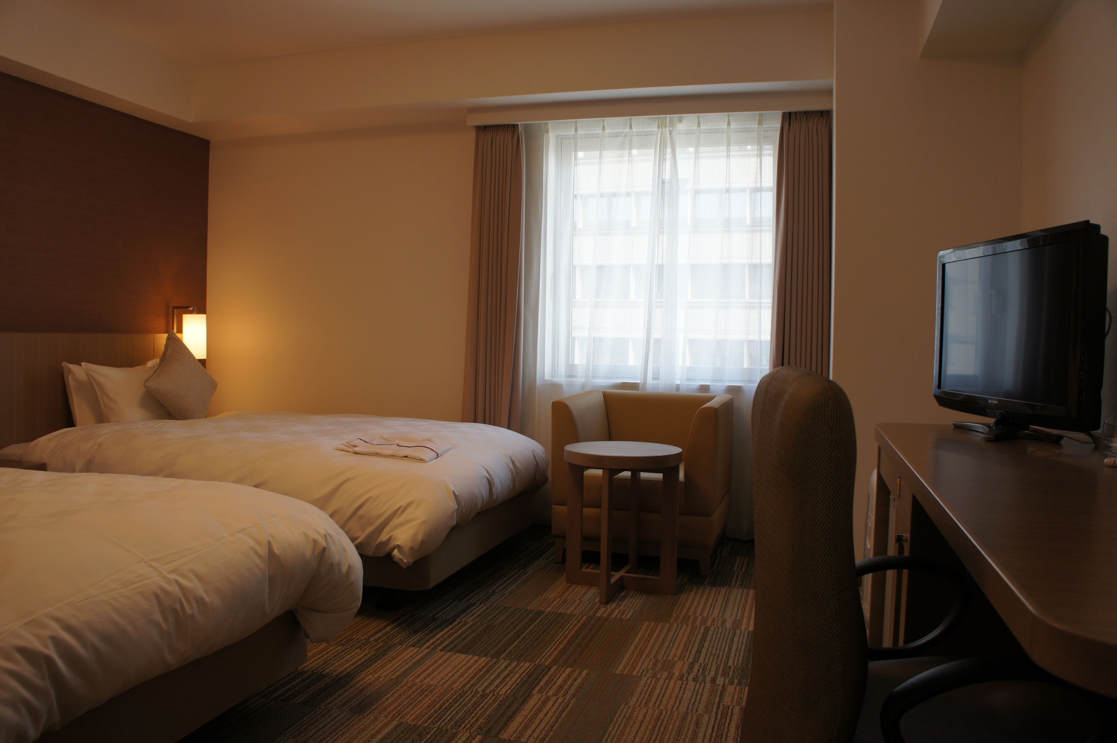 A hotel room with two beds, a small table, a chair, a TV, and a window with curtains.