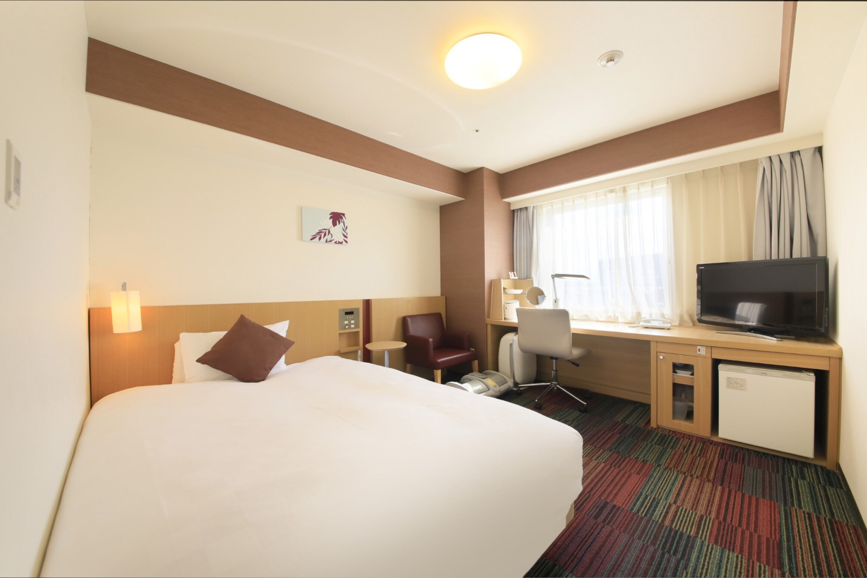 A hotel room with a large bed, a desk with a television, a chair, and a small refrigerator.