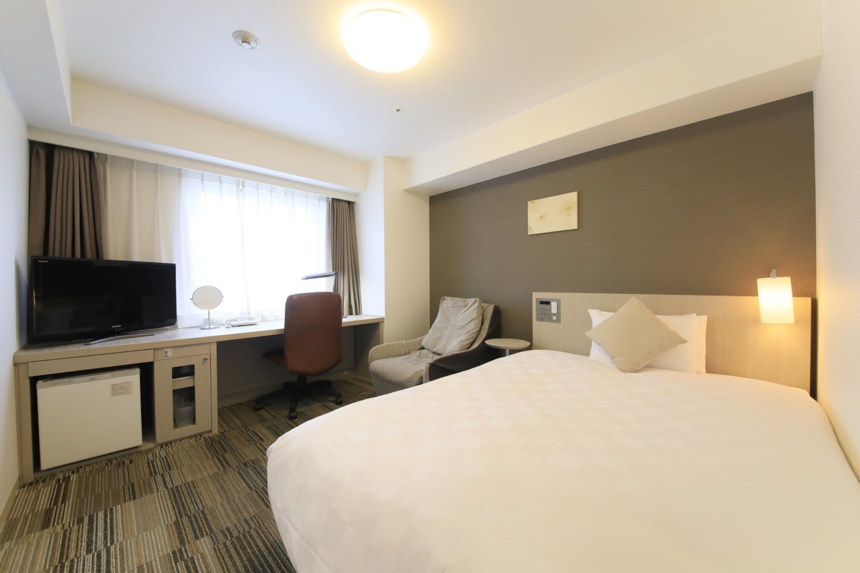 Quarto de hotel com cama, escrivaninha com televisão, cadeira, secretária pequena e janela com cortinas.