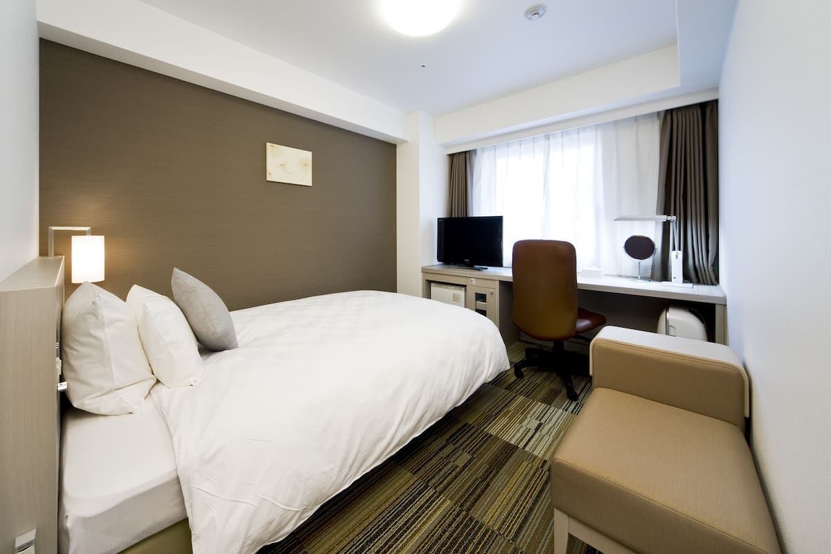 Hotelkamer met een bed, een bureau met een computer, een stoel en een raam met gordijnen.