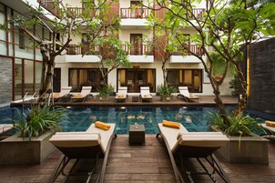 Uma área à beira da piscina com espreguiçadeiras e uma mesa pequena, cercada por plantas em vasos e um edifício com varandas.