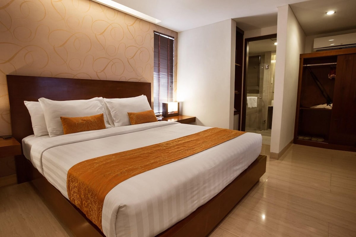 A hotel room with a large bed, a wooden headboard, a nightstand, a bedside table, a bathroom with a glass shower, and a walk-in closet.