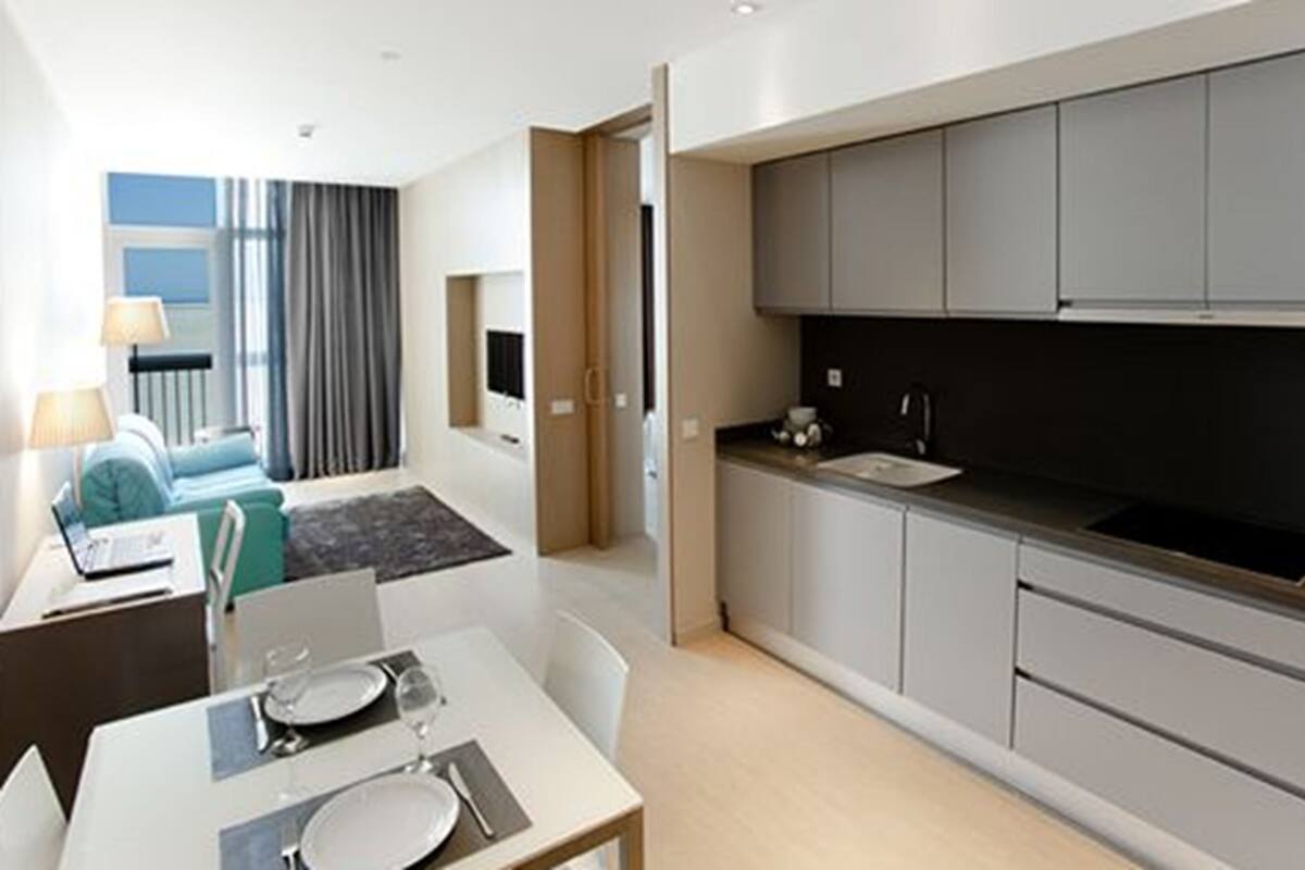 Eine moderne Küche mit grauen Schränken, einer schwarzen Arbeitsplatte und einem Spülbecken. Es gibt einen Essbereich mit einem für zwei Personen gedecktem Tisch sowie eine Wohnbereicht mit einer Couch und einer Lampe.