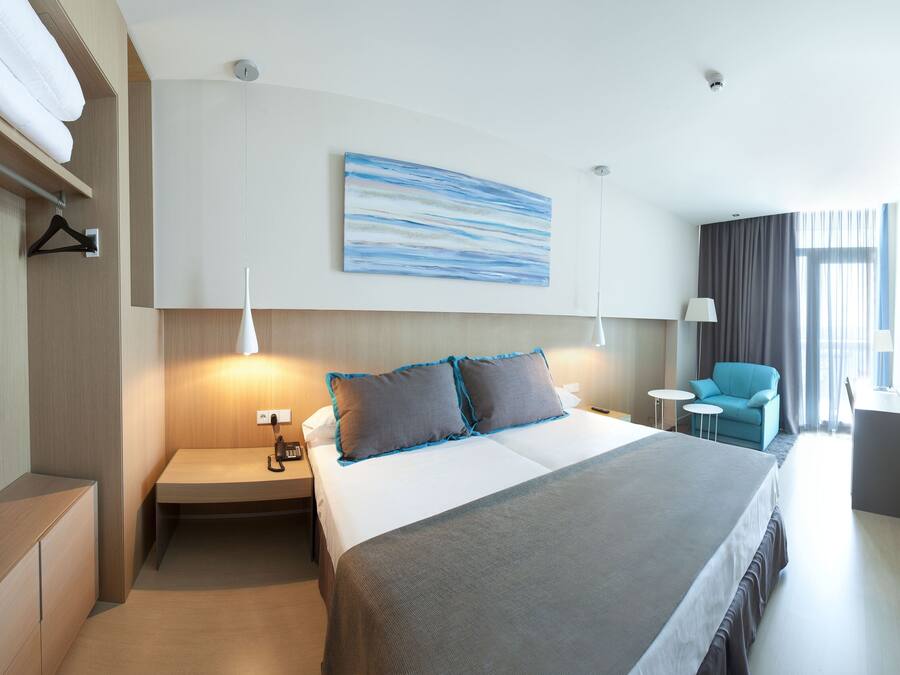 A modern hotel room with a large bed, bedside tables, a desk, a chair, and a painting on the wall.