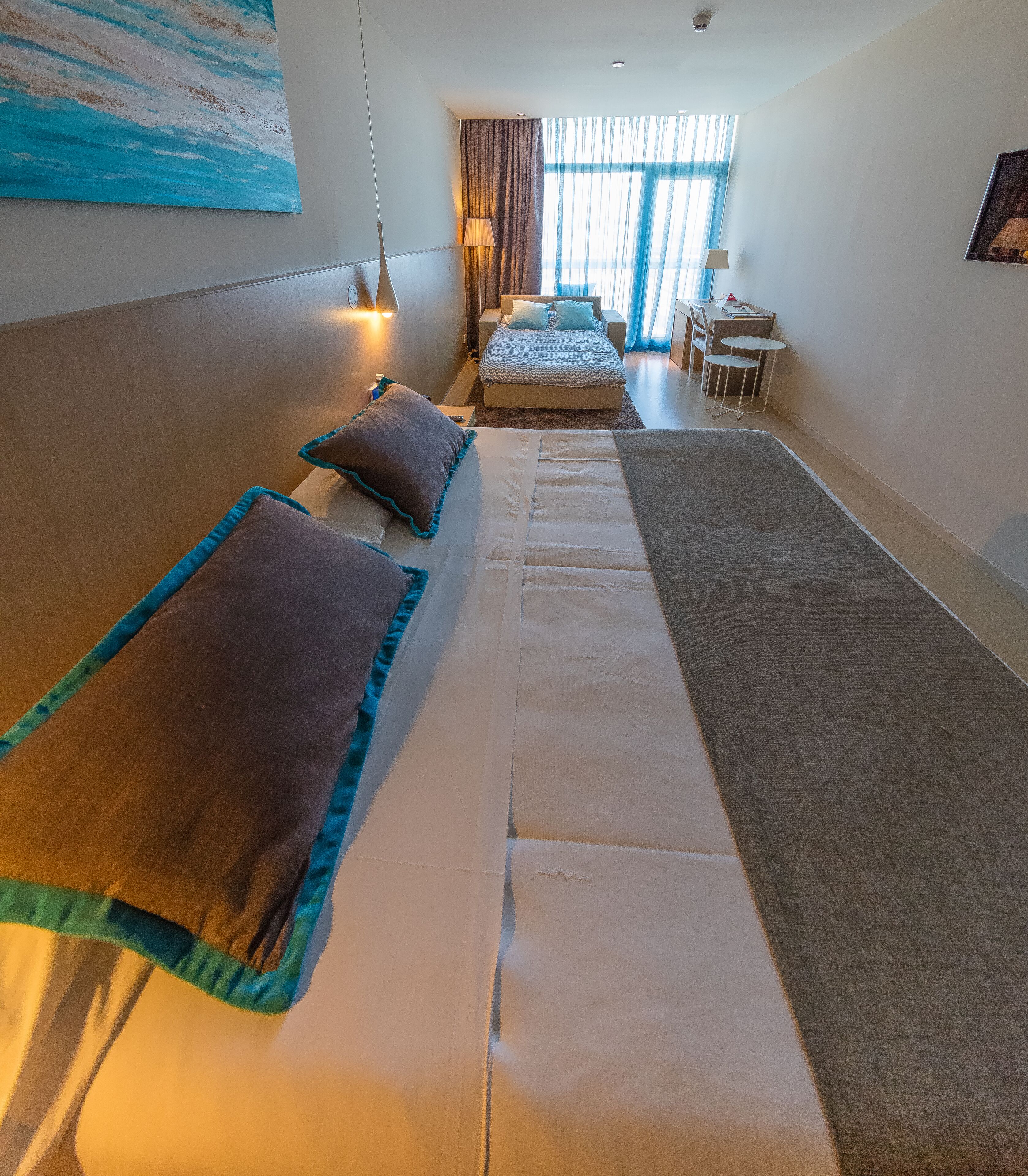 Hotellihuone, jossa on pitkä sänky, pieni pöytä ja tuoli.