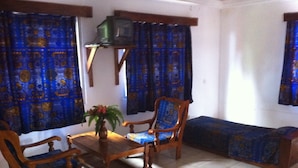 A living room with a wooden sofa, two wooden chairs, a small table with a vase, and blue patterned curtains. - Napoléon Aéroport (Lomé)