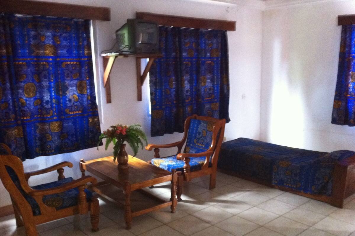Una sala de estar con un sofá de madera, dos sillas de madera, una mesita con un florero y cortinas azules estampadas.