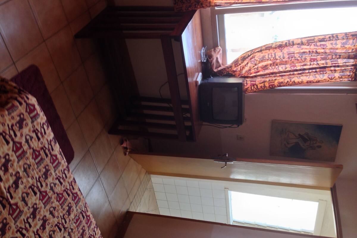Una habitación con suelo de baldosas, una escalera de madera, un televisor sobre un mueble bajo y un ventanal con cortinas estampadas.
