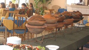 A buffet setup with multiple large pots and bowls on a table, surrounded by chairs and tables with blue tablecloths.