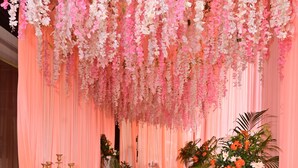 Un pasillo con cortinas rosas, adornos florales colgantes, plantas en macetas y un candelabro.