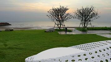 A grassy outdoor area with a white, perforated table in the foreground, two leafless trees, and a view of the sea in the background.