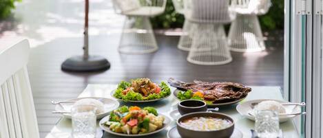 Una mesa de comedor con diversos platos, rodeada de sillas blancas, ubicada en una terraza con vistas a la vegetación y una piscina.