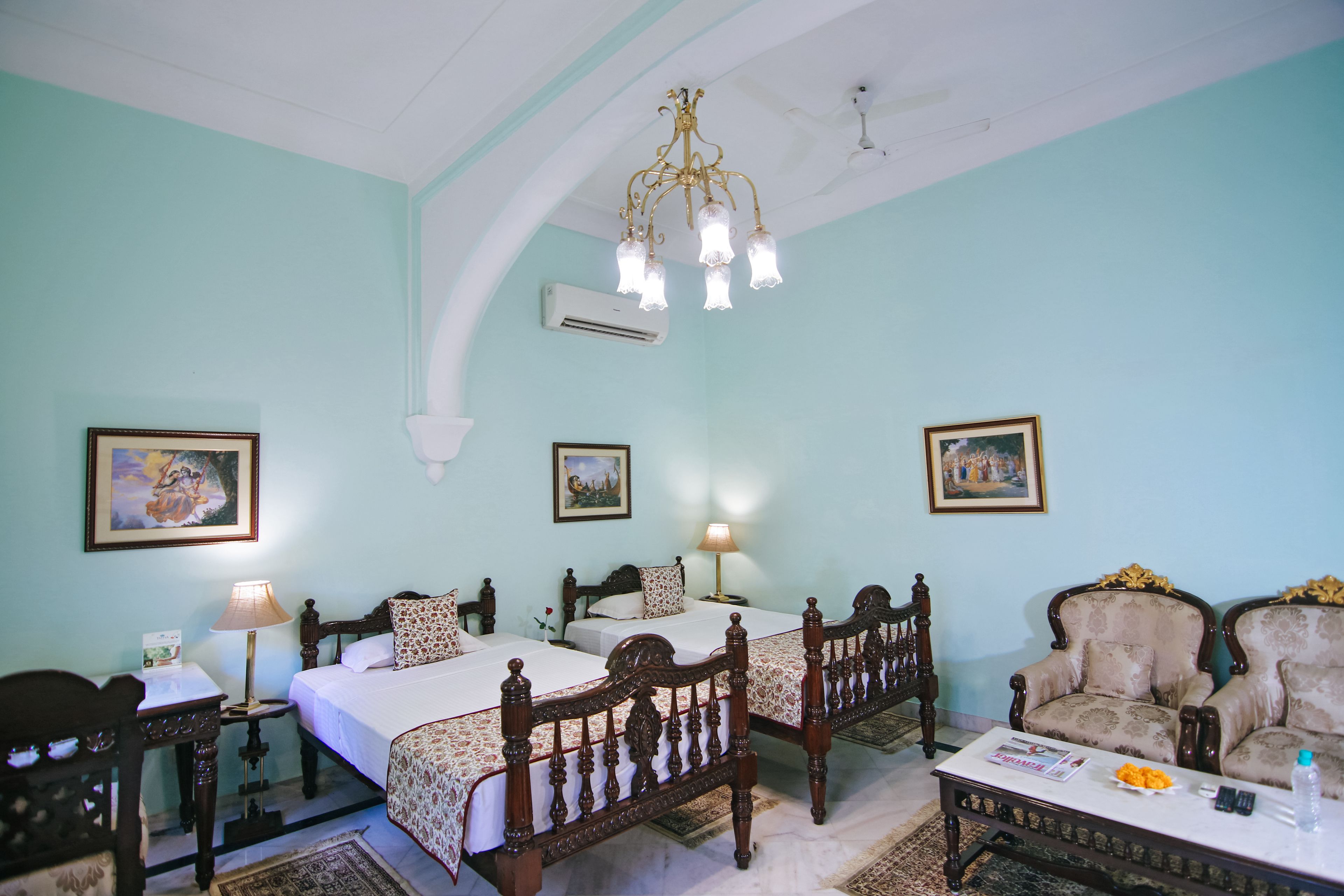 A room with two beds, a wooden chair, a table with a magazine, and a chandelier.