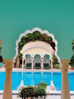Zona de piscina con fuente, sillas reclinables verdes y un arco decorativo.