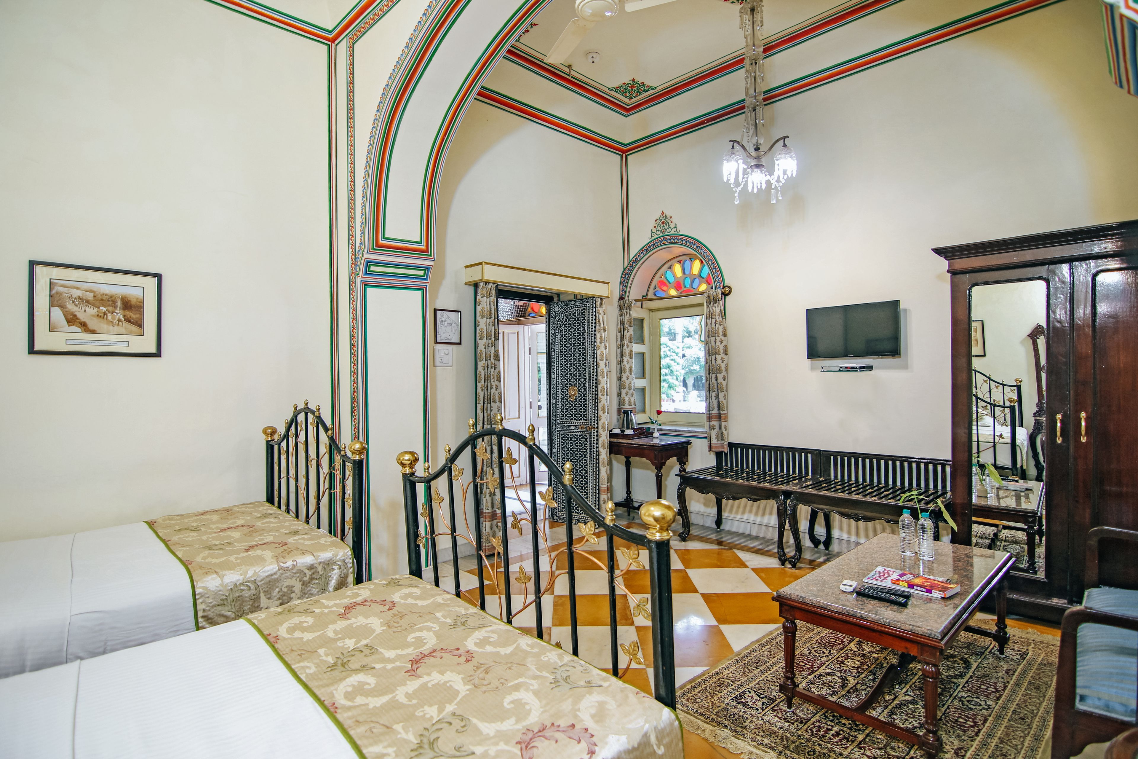 A room with two beds, a wooden bench, a table with a rug, a television, and a chandelier.