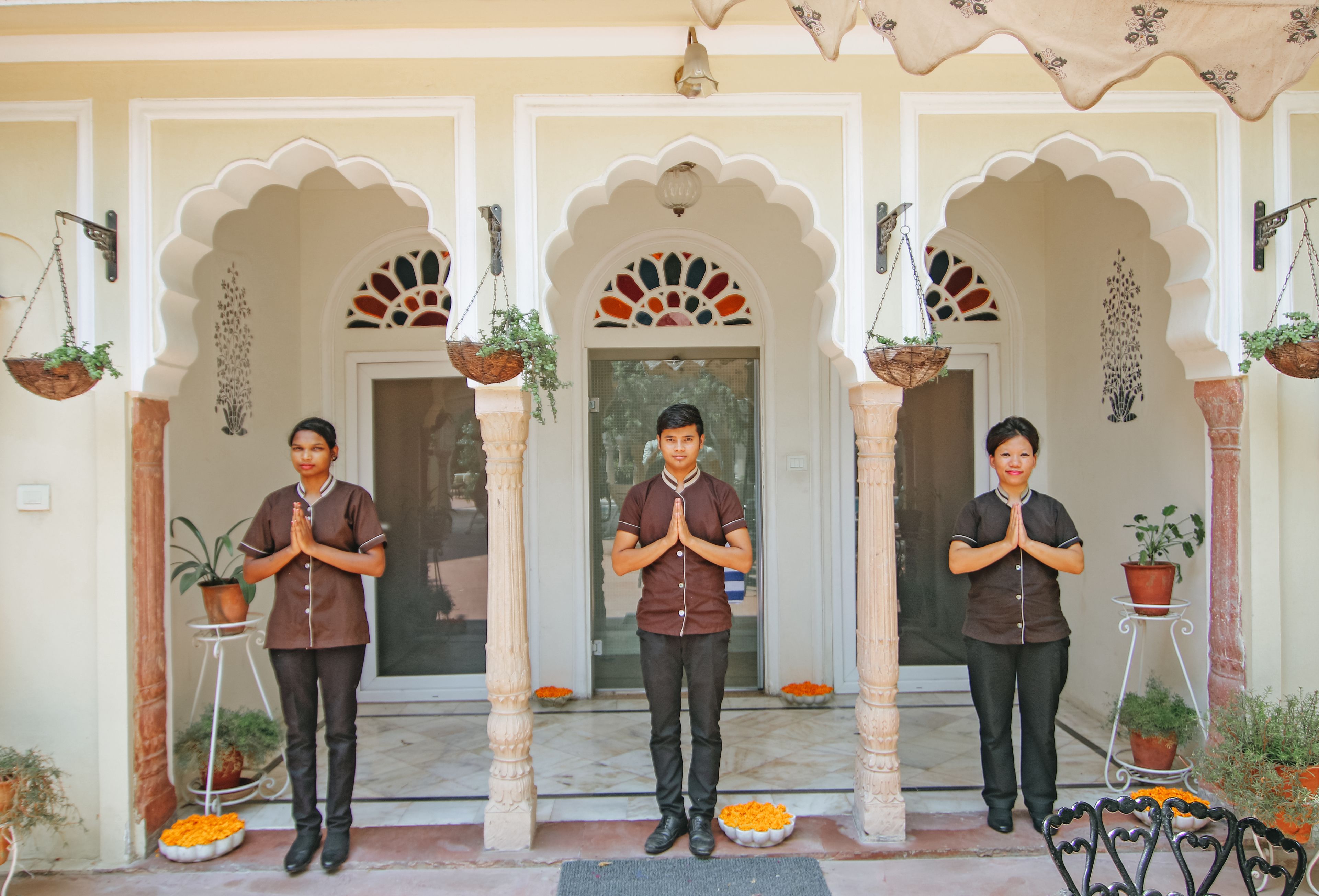 Three individuals standing in a doorway with decorative arches, potted plants, and orange flowers on the ground.