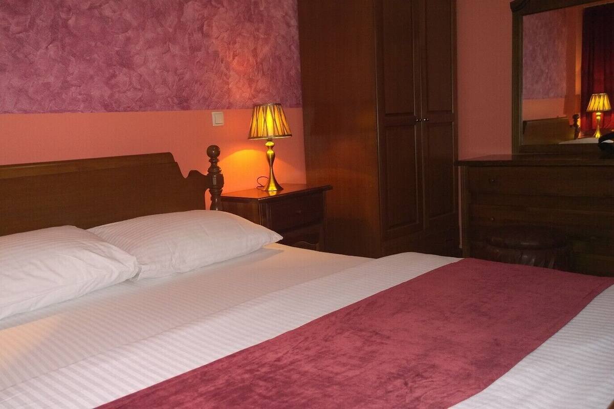 A neatly made bed with white linens and a pink blanket, a wooden headboard, a bedside lamp, and a wooden wardrobe.