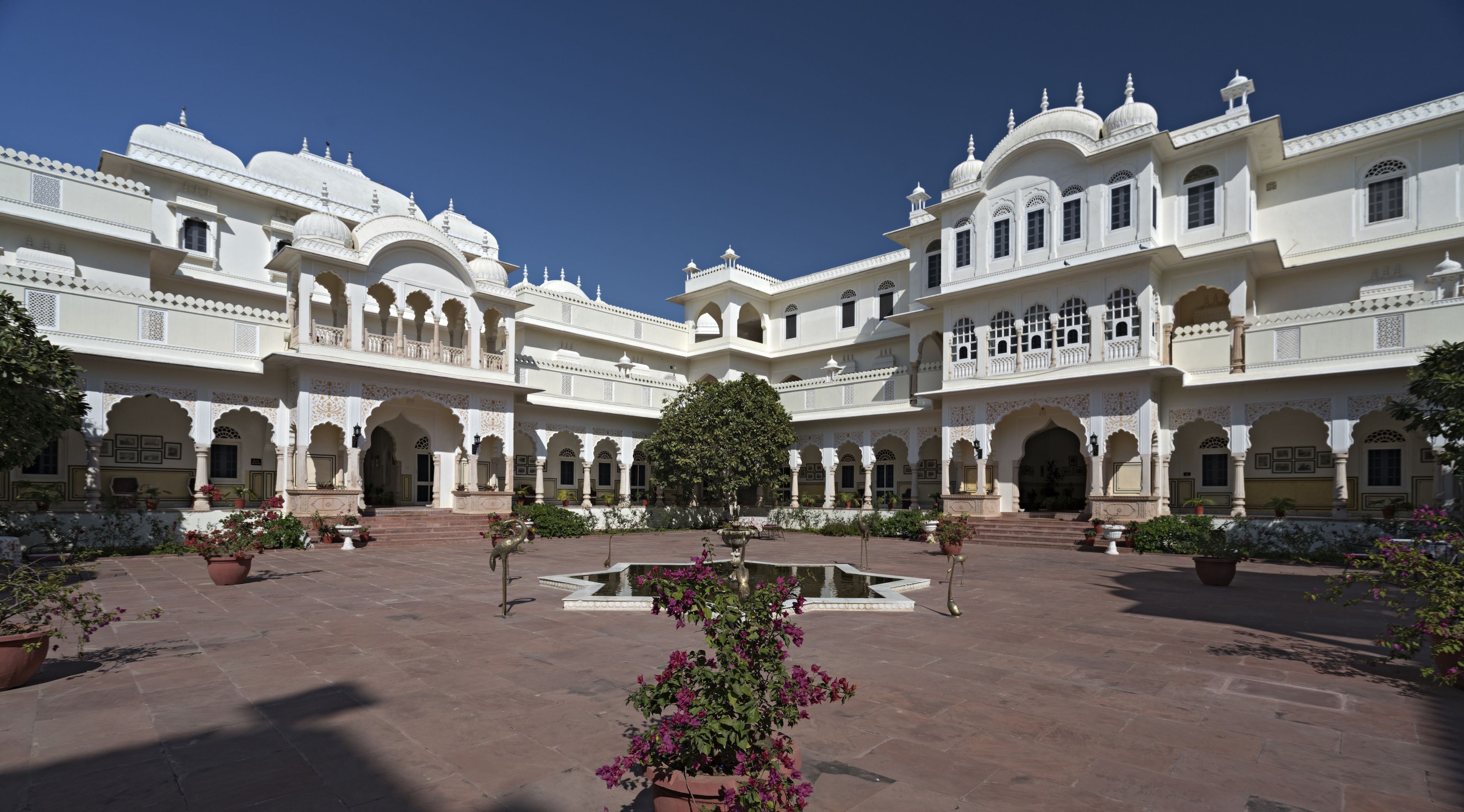 A large, white, multi-story building with arched windows and balconies, surrounded by a courtyard with a central water feature and potted plants.