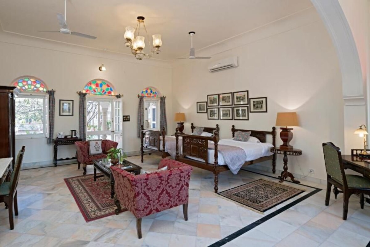 A spacious room with a large bed, a dining table, and chairs. There are two windows with stained glass, a chandelier, and framed pictures on the wall.