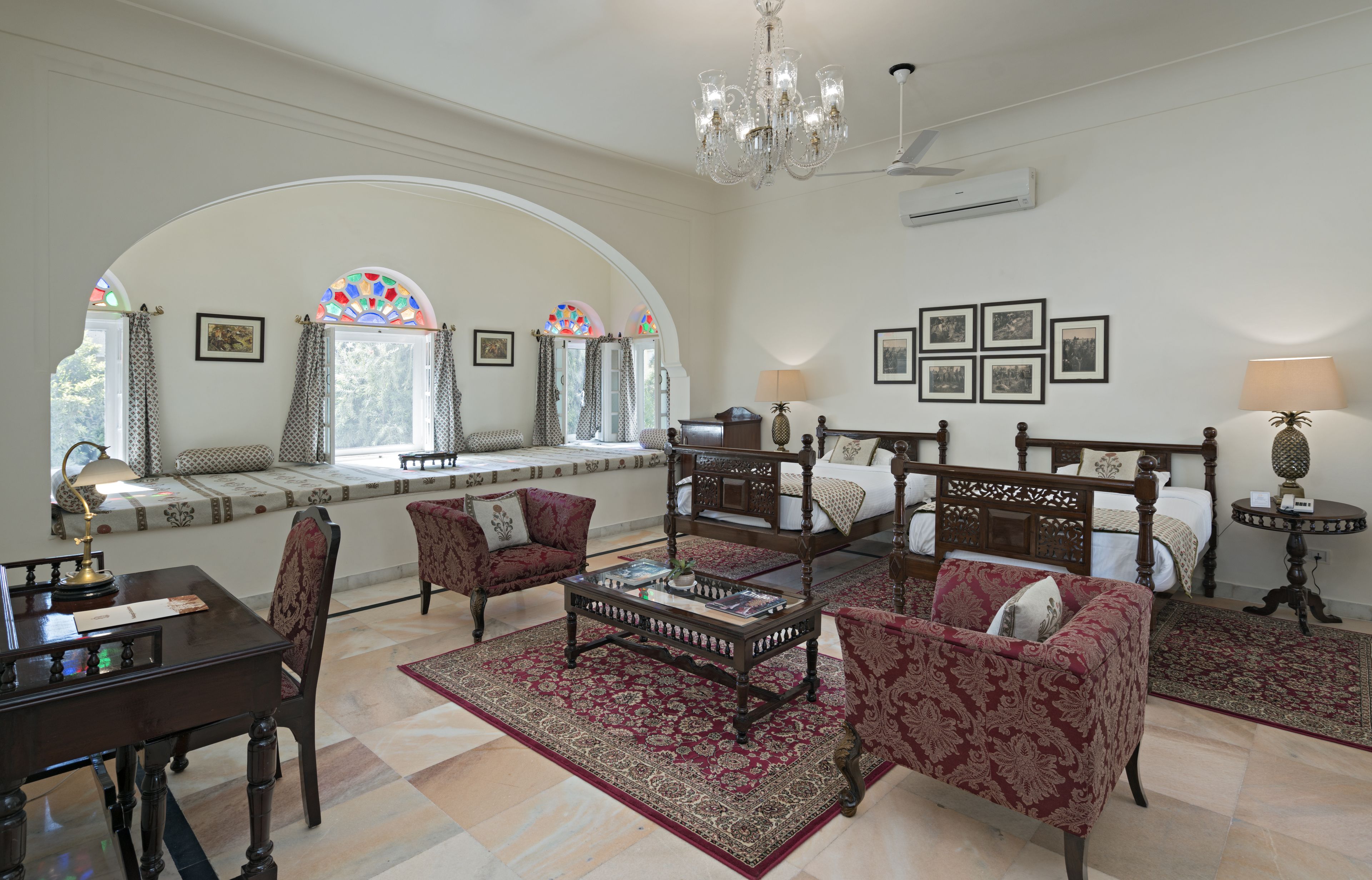 A spacious living room with a large bed, a dining table, and a coffee table. There are multiple windows with stained glass, a chandelier, and framed pictures on the walls.