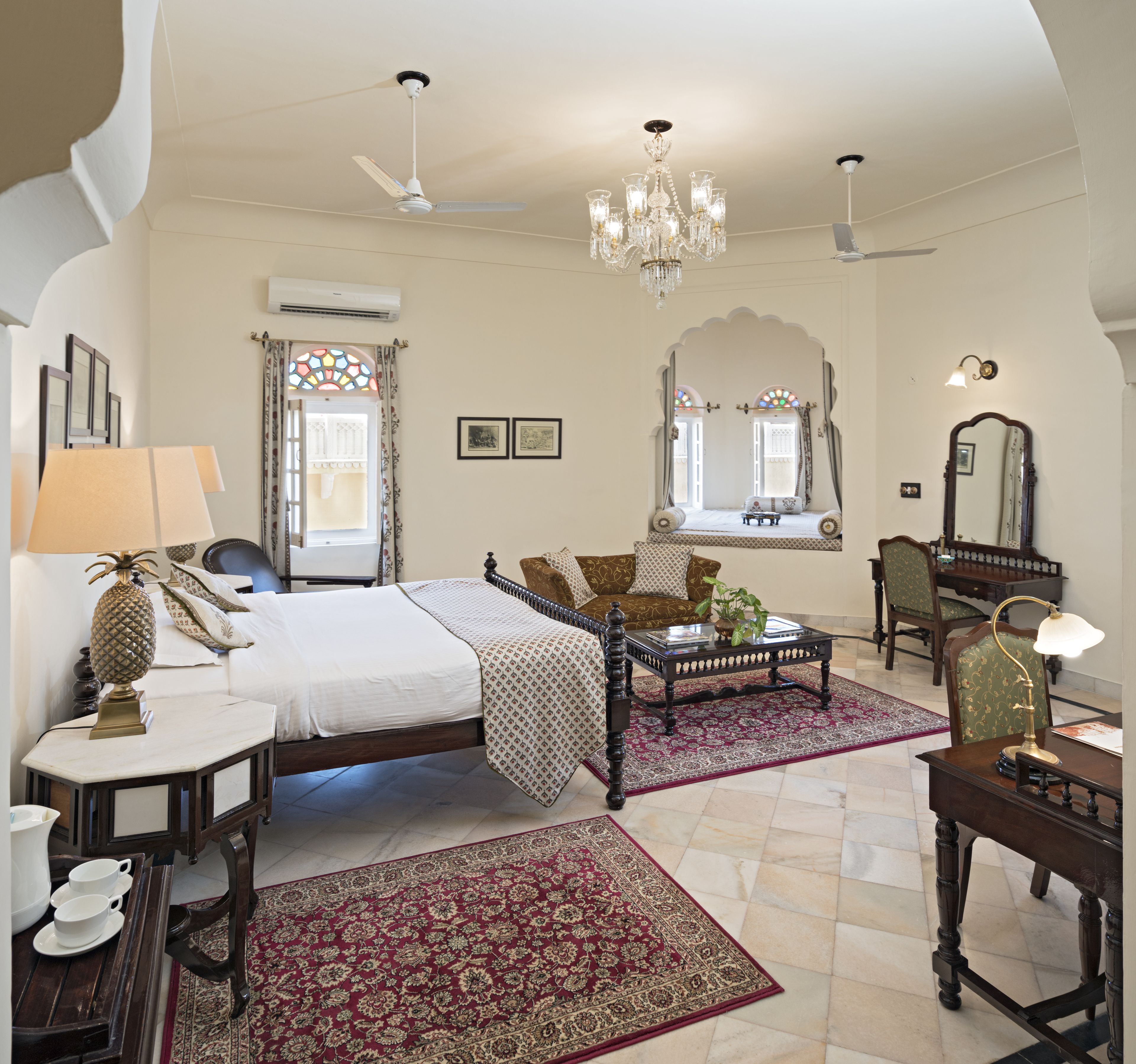 A spacious bedroom with a large bed, a sitting area with a sofa and armchairs, a chandelier, and a large mirror.