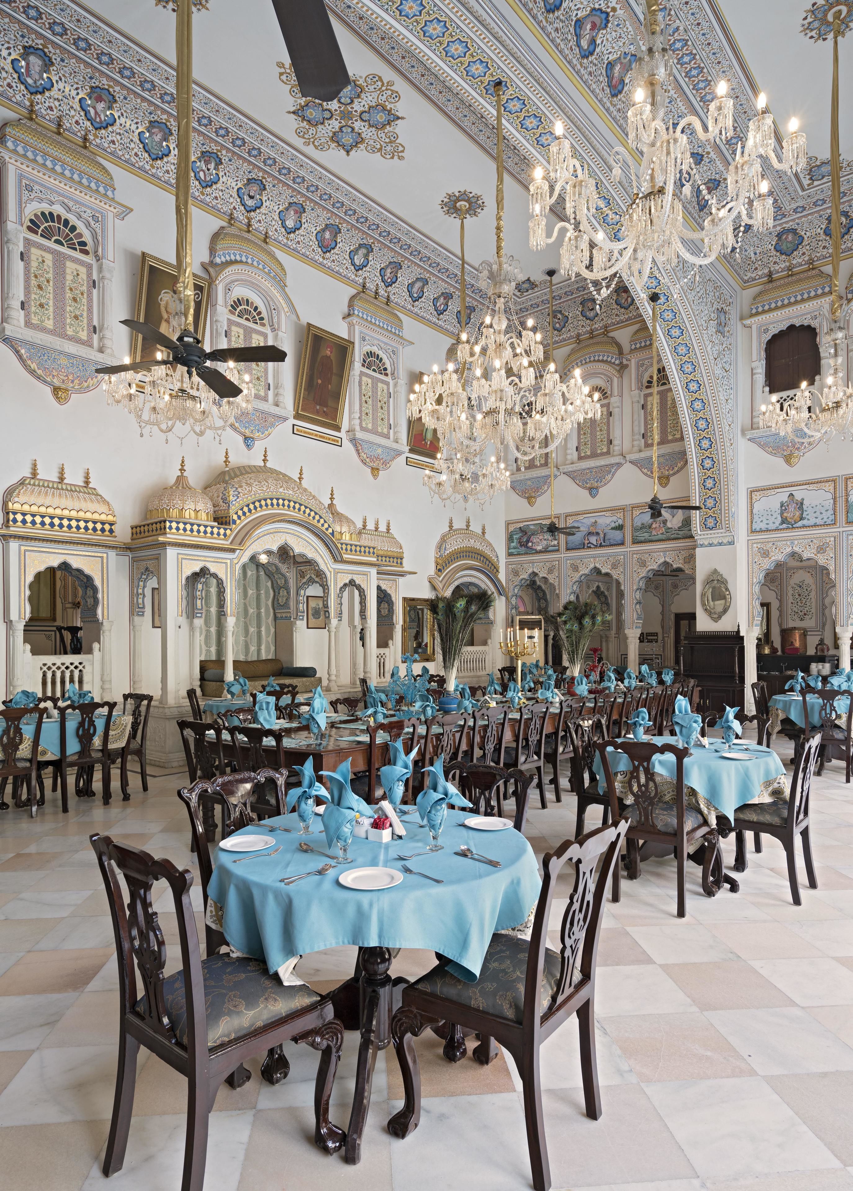 Un vaste réfectoire agrémenté de lustres somptueux, de hauts plafonds et de décorations murales complexes. Les tables sont dressées avec des nappes bleues et de l’argenterie.