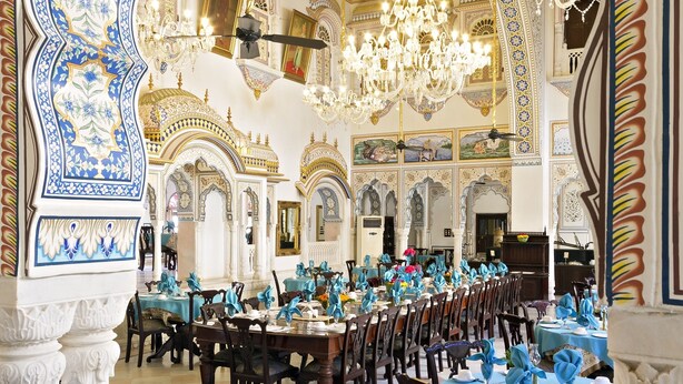 Un salón comedor amplio con diseños ornamentales en el techo, azul y blanco, candelabros y un ventilador de techo. Las mesas están dispuestas con manteles azules y centros de mesa dorados.