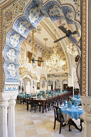 A grand dining hall with ornate blue and white ceiling designs, chandeliers, and a ceiling fan. Tables are set with blue tablecloths and gold centerpieces. - Alsisar Mahal - A Heritage Hotel (Malasisar)