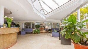 A spacious lobby with a high ceiling and skylight, featuring seating areas, a reception desk, and potted plants.
