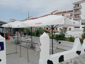 An outdoor seating area with white umbrellas labeled "VICTORIA," featuring tables and chairs, surrounded by buildings with balconies.