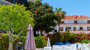 A poolside area with white lounge chairs and a purple umbrella, surrounded by lush greenery and a building with a red-tiled roof.