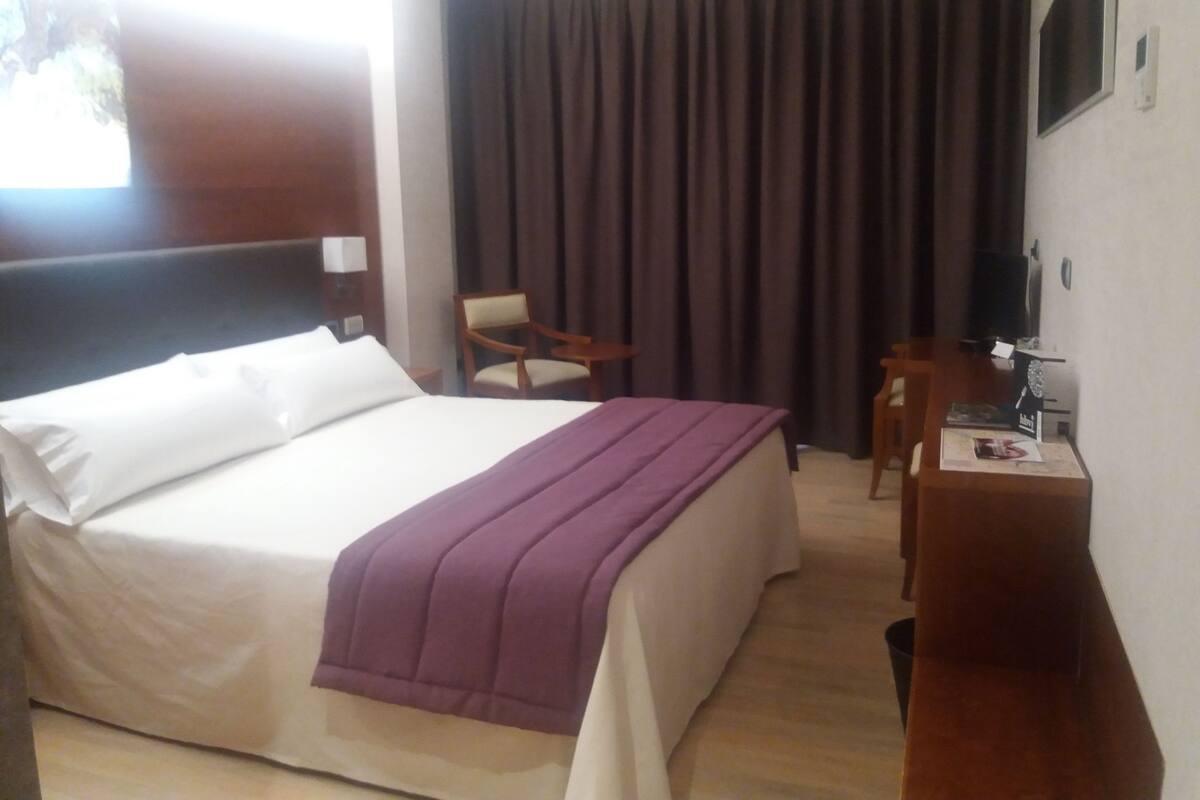 A hotel room with a bed, a desk, a chair, and a large window with curtains.