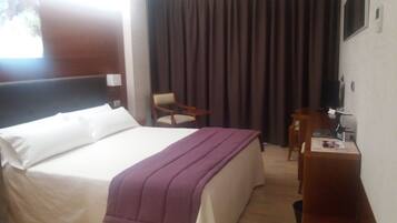 Habitación de hotel con cama, escritorio, silla y una ventana grande con cortinas.