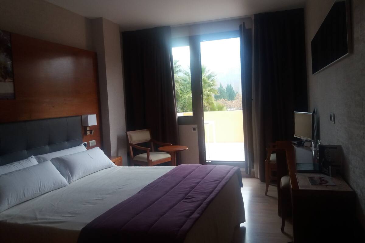 A hotel room with a bed, a desk with a computer, a chair, and a view of the outdoors.