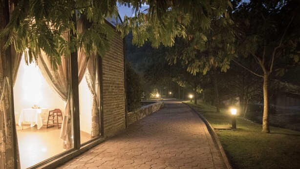 A paved pathway leading to a building with a large window and curtains, a brick wall, and a grassy area with trees. - Balneario Valle del Jerte (Valdastillas)