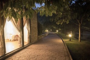 Un camino pavimentado que conduce a un edificio con un amplio ventanal y cortinas, un muro de ladrillo y una zona verde con árboles.
