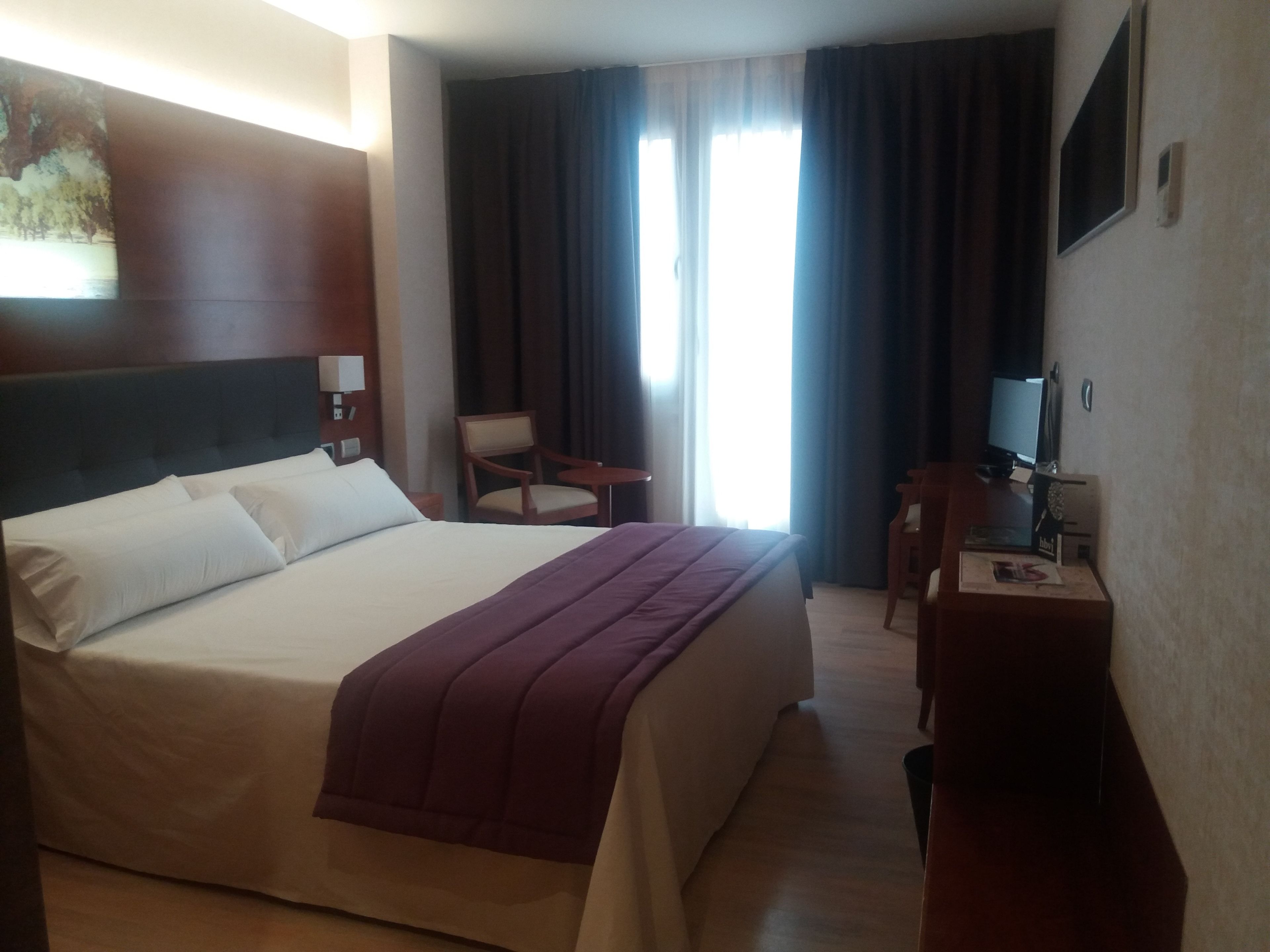 Habitación de hotel con una cama grande, un escritorio, una silla, un televisor y una ventana con cortinas.