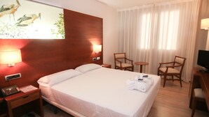 A hotel room with a large bed, two bedside tables, a desk with a chair, a small round table with a chair, and a television. - Balneario Valle del Jerte (Valdastillas)
