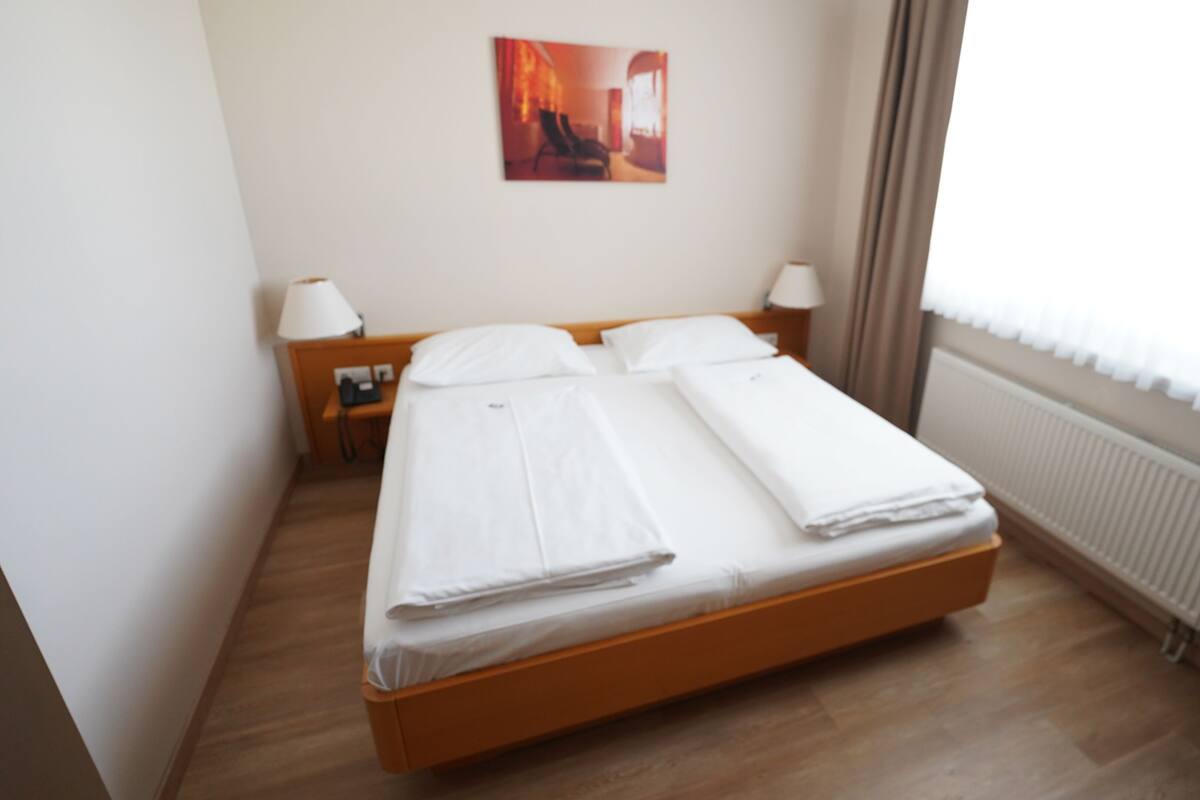 Una cama bien hecha con sábanas blancas en una habitación de hotel. Se ve un mesita de noche con una lámpara y un teléfono. Un cuadro cuelga en la pared sobre la cama.