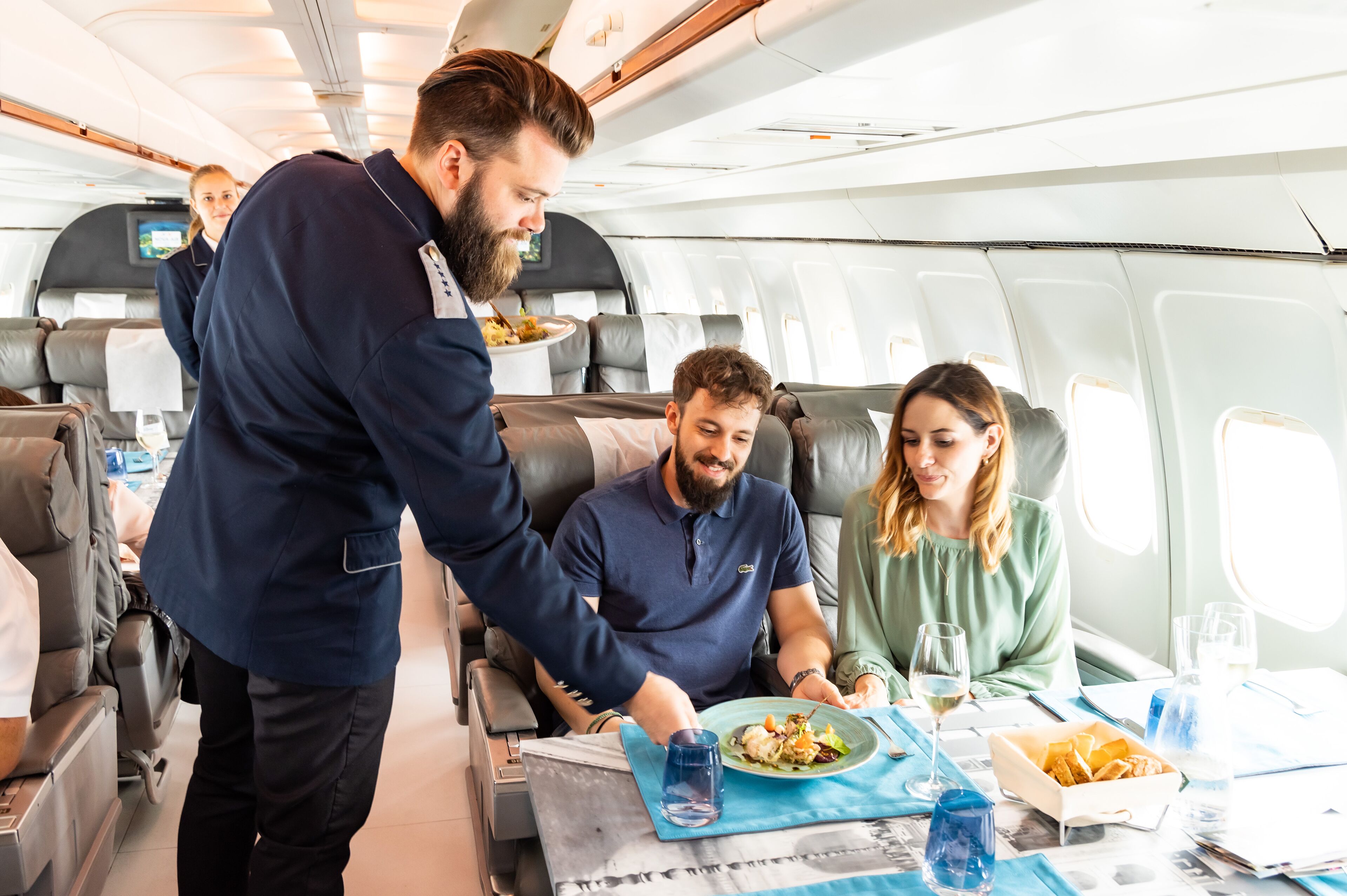 Ein Flugzeuginnenraum mit Essensbrett, sitzenden Passagieren und einem Flugbegleiter, der Speisen serviert.
