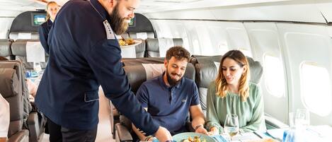 Ein Flugzeuginnenraum mit Essensbrett, sitzenden Passagieren und einem Flugbegleiter, der Speisen serviert.